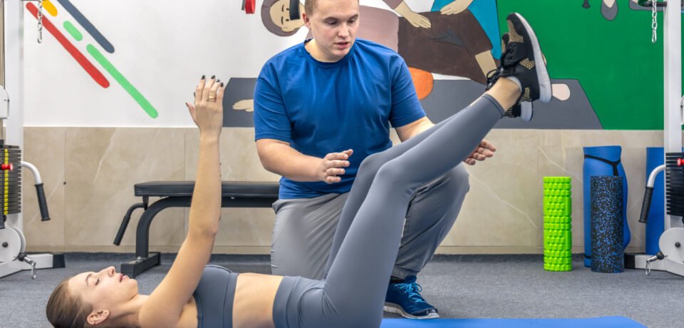 In a rehabilitation clinic a patient practices a core move with legs raised on a blue mat while a doctor cues breathing and control. Calm wellness mood supports mobility and recovery content.